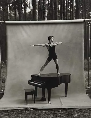 Photographie en noir et blanc d'un danseur athlétique en justaucorps noir, posant en équilibre sur un piano à queue installé en pleine forêt devant un fond de studio.