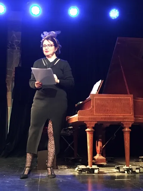 Sabine Miniconi sur scène sous les projecteurs, lisant un texte poétique devant un piano à queue lors d'un spectacle piano et poésie